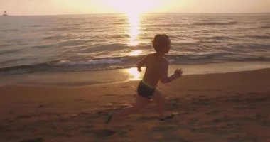 Gün batımında sahilde koşan bir çocuk. Genç çocuk kum ve denizde eğleniyor. Arka planda parlayan altın güneş