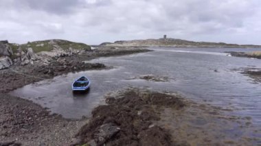 Dalgaların alçak deniz manzarası. Kayalardan ve yosunlardan oluşan alçak sularda mavi bir tekne. Düşük gelgitte, arka plandaki deniz fenerinde, İrlanda kıyı şeridinin insansız hava aracı bakış açısı