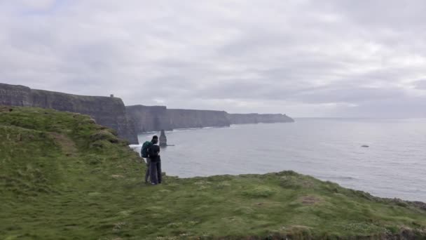 Widok z lotu ptaka na piękne irlandzkie klify, młoda para ogląda krajobraz, panoramę. Chmurne niebo w tle, wzburzone morze. Słynne miejsce w Irlandii, Matka klifów — Wideo stockowe