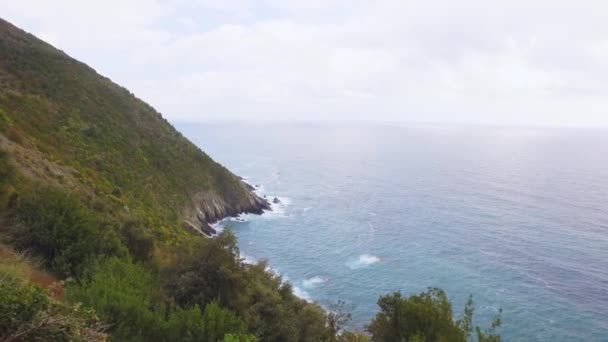 Περπατώντας σε χωματόδρομο, δίπλα στη γαλάζια θάλασσα στο Cinque Terre. Ορεινή πλαγιά καλυμμένη από θάμνους και θαλάσσια δέντρα. Γαλήνιος καιρός με σύννεφα. Κύματα σπάνε πάνω από τους βράχους — Αρχείο Βίντεο