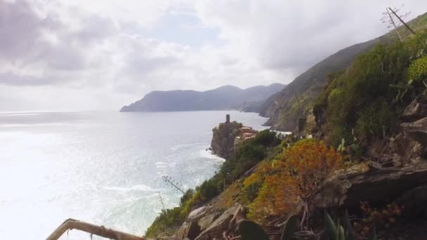 Berjalan melalui jalur tanah, di samping laut biru di Cinque Terre, di jarak Vernazza. Lereng gunung ditutupi semak-semak dan pohon maritim. Cuaca tenang dengan beberapa awan. Gelombang melanggar bebatuan — Stok Video