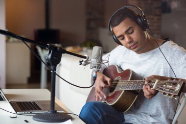 Afro-Amerikalı genç erkek müzisyen sandalyede oturup gitar çalma alıştırması yapıyor. Evde mikrofonlu profesyonel müzik ekipmanları ve dizüstü bilgisayar dersi kullanıyor.