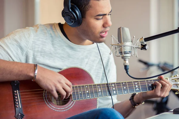 Giovane musicista afro-americano seduto e che canta sulla sedia esercitandosi a suonare la chitarra usando il suo portatile, attrezzatura musicale professionale con microfono e cuffia a casa Foto Stock