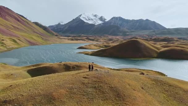 Flygfoto av en vacker dal med omgivande berg, två personer på toppen av backen. Floden korsade grön och brun dal under solig dag.Tagikistan — Stockvideo