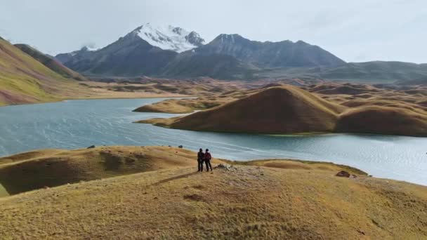 Εναέρια λήψη μιας όμορφης κοιλάδας με γύρω βουνά, δύο άνθρωποι στην κορυφή του λόφου. Ποταμός διέσχισε πράσινο και καφέ κοιλάδα κατά τη διάρκεια της ηλιόλουστης ημέρας.Tagikistan — Αρχείο Βίντεο