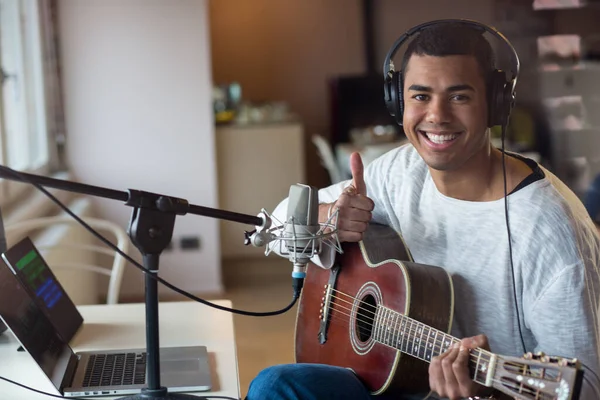 Afro american young man male musician sitting and smiling on chair practicing learning playing guitar using his laptop, professional music equipment with microphone and headphone at home Stock Image