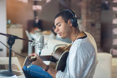 Afro-Amerikalı genç erkek müzisyen sandalyede oturup gitar çalma alıştırması yapıyor. Evde mikrofonlu profesyonel müzik ekipmanları ve dizüstü bilgisayar dersi kullanıyor.