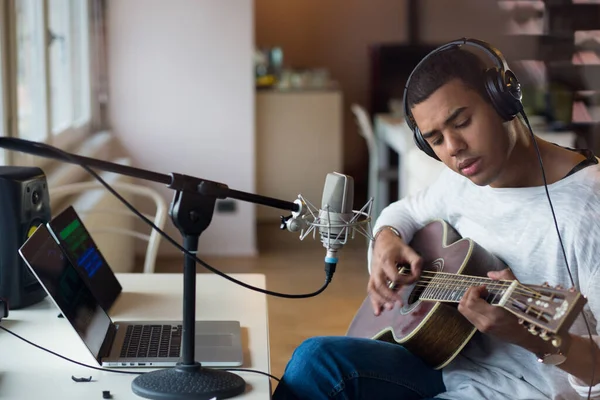 Afro american young man male student musician sitting on chair practicing learning playing guitar using online teacher video lesson in laptop, professional music equipment with microphone at home Stock Photo