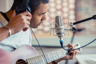 Afro-Amerikalı genç erkek müzisyen sandalyede oturup gitar çalma pratiği yapıyor. Evde mikrofon ve kulaklıkla müzik yapıyor.
