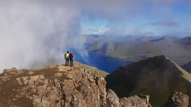 Вид с воздуха на двух человек на вершине горы с видом на красивый пейзаж, море, скалу, гору, скалу, обертывание облаками — стоковое видео