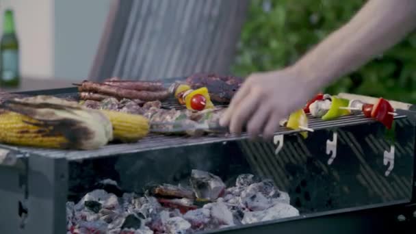 Pimientos, chiles verdes, maíz, cebolla, hamburguesas, mocosos y cabritos dispuestos en una parrilla de barbacoa. Primer plano del joven cocinando carne para la barbacoa. — Vídeos de Stock