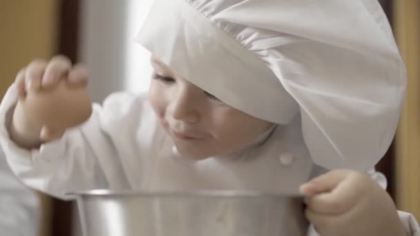 Nahaufnahme der Hände eines jungen Kochs, der lernt, Eier zu brechen, Hobby. Zutaten zur Zubereitung von Kuchen. Sohn in weißer Koch-Uniform backt in seiner Küche zu Hause mit seinem Vater eine Torte. Zeitlupe. — Stockvideo