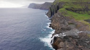 Faroe Adaları 'ndaki kaya uçurumunun havadan görünüşü. Mavi deniz dalgaları, bulutlu yolculukta dalgalı deniz. yavaş çekim