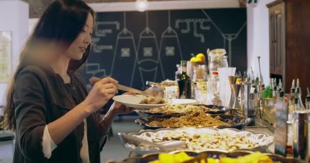 Jovem mulher oriental selecionando comida no restaurante self-service. Mulher bonita na frente de uma mesa de comida coberta auto-atendimento. — Vídeo de Stock