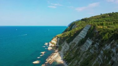 Mavi denizin aşağısındaki kayalık kıyı şeridinin havadan görünüşü. Beyaz kayalar, ağaçlar. Arka planda hareket eden bir tekne, İtalya, Conero 'da bir yaz gününde mavi gökyüzü