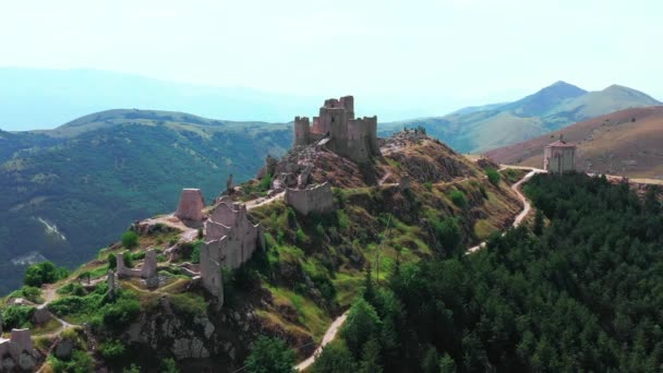 Vista aérea del antiguo castillo en la colina de la montaña, bosque de pinos en la ladera de la montaña.Rocas blancas y camino sucio curvado a lo largo de la montaña. Cordillera en el fondo con cielo azul un día soleado de verano — Vídeo de stock