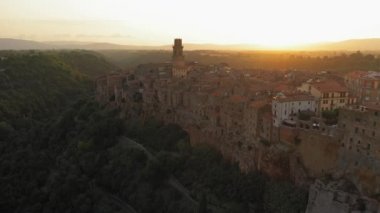 Günbatımında gökyüzü manzaralı antik binalar, Pitigliano, Toskana. Ortaçağ mimarisi dağ tepesinde, çam ormanı boyunca kıvrımlı yol. Evlerde güneş parlıyor