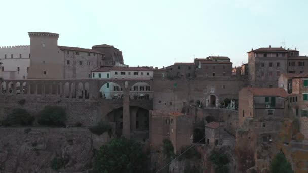 Vista frontal aérea de edificios antiguos durante la puesta del sol, Pitigliano, Toscana. Arquitectura medieval o colina de montaña, camino curvo a través del bosque de pinos. Sol brillando a través de las casas — Vídeos de Stock