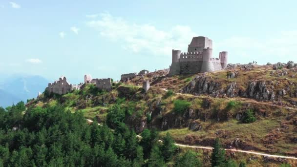 Widok z lotu ptaka starożytnego zamku na wzgórzu, sosnowy las na zboczu góry.Białe skały i zakrzywiona brudna droga wzdłuż góry. Pasmo górskie w tle z błękitnym niebem un letni słoneczny dzień — Wideo stockowe