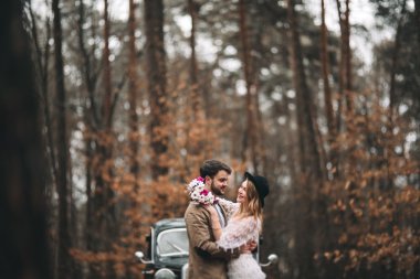 Şık Loving düğün çift öpüşme ve retro car yakınındaki bir çam ormanı içinde sarılma