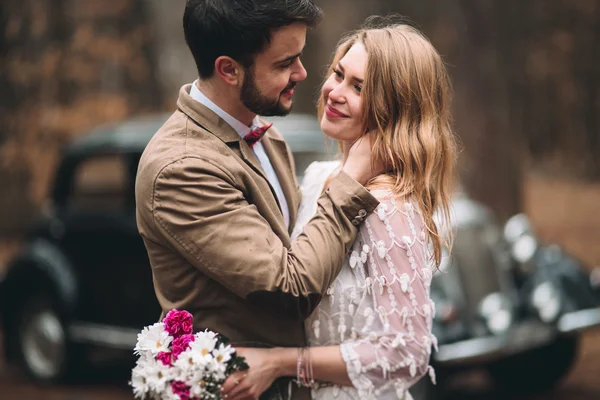 Elegante pareja de boda cariñosa besándose y abrazándose en un bosque de pinos cerca de coche retro —  Fotos de Stock