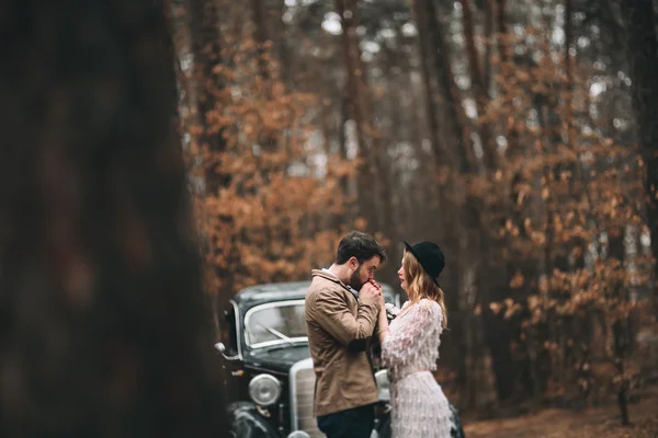 Стильная влюбленная свадебная пара целуется и обнимается в сосновом лесу рядом с ретро-машиной — стоковое фото