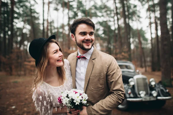 Elegante pareja de boda cariñosa besándose y abrazándose en un bosque de pinos cerca de coche retro —  Fotos de Stock