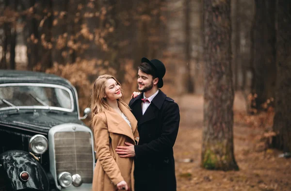 Stylish Loving wedding couple kissing and hugging in a pine forest near retro car — Stock Photo, Image