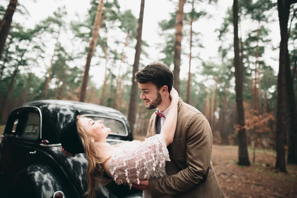 Casal amoroso elegante beijando e abraçando em uma floresta de pinheiros perto de carro retro — Fotografia de Stock