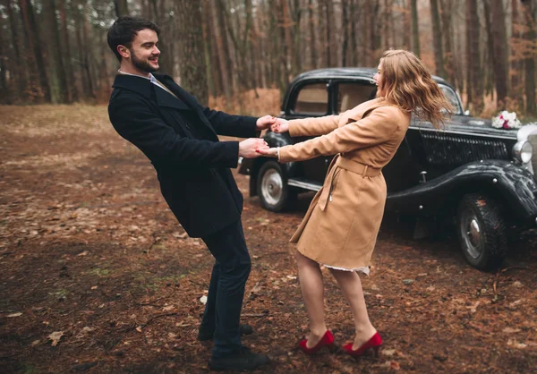 Elegante pareja de boda cariñosa besándose y abrazándose en un bosque de pinos cerca de coche retro — Foto de Stock