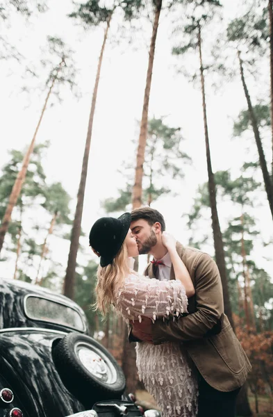 Şık Loving düğün çift öpüşme ve retro car yakınındaki bir çam ormanı içinde sarılma