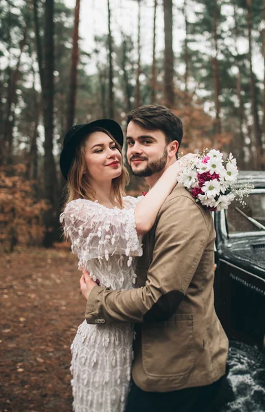 Casal amoroso elegante beijando e abraçando em uma floresta de pinheiros perto de carro retro — Fotografia de Stock