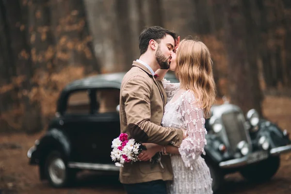Elegante pareja de boda cariñosa besándose y abrazándose en un bosque de pinos cerca de coche retro —  Fotos de Stock