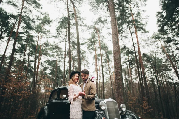 Stylish Loving wedding couple kissing and hugging in a pine forest near retro car — Stock Photo, Image