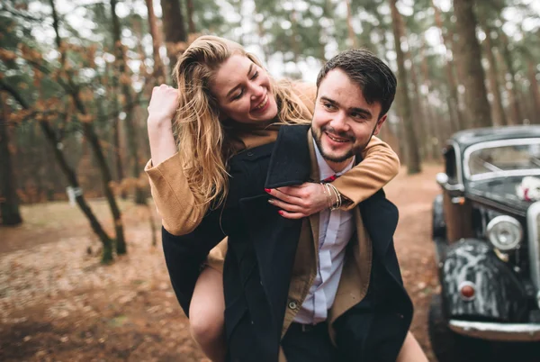 Elegante pareja de boda cariñosa besándose y abrazándose en un bosque de pinos cerca de coche retro — Foto de Stock