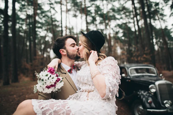 Stylish Loving wedding couple kissing and hugging in a pine forest near retro car — Stock Photo, Image