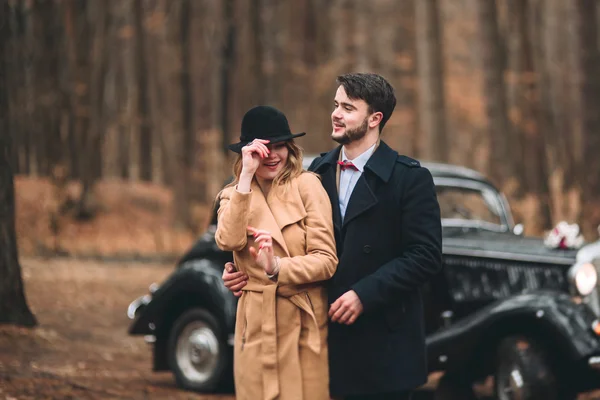 Stylish Loving wedding couple kissing and hugging in a pine forest near retro car — Stock Photo, Image