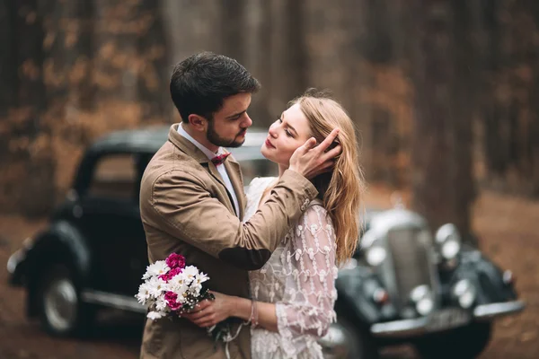 Şık Loving düğün çift öpüşme ve retro car yakınındaki bir çam ormanı içinde sarılma