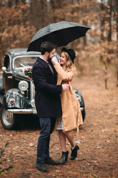 Stílusos szerető esküvői pár csók és egy fenyves erdő közelében retro autó átölelve — Stock Fotó