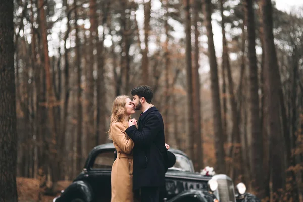 Casal amoroso elegante beijando e abraçando em uma floresta de pinheiros perto de carro retro — Fotografia de Stock