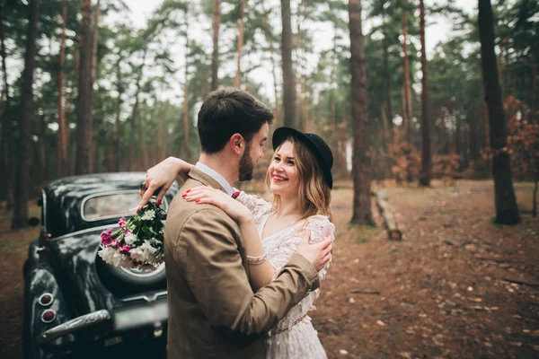 Стильная влюбленная свадебная пара целуется и обнимается в сосновом лесу рядом с ретро-машиной — стоковое фото