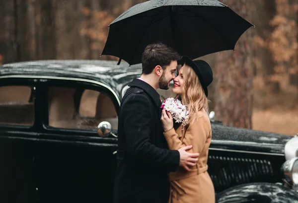 Casal amoroso elegante beijando e abraçando em uma floresta de pinheiros perto de carro retro — Fotografia de Stock