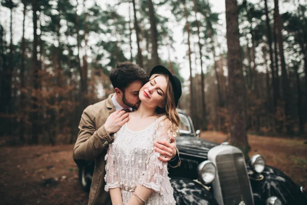 Stijlvolle Loving bruiloft paar zoenen en knuffelen in een dennenbos in de buurt van retro auto — Stockfoto