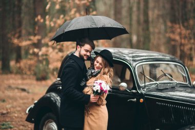 Gorgeous newlywed bride and groom posing in pine forest near retro car in their wedding day