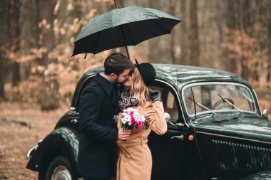 Gorgeous newlywed bride and groom posing in pine forest near retro car in their wedding day
