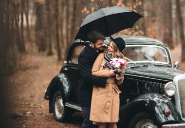 Gorgeous newlywed bride and groom posing in pine forest near retro car in their wedding day