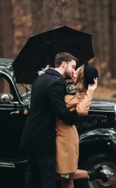 Gorgeous newlywed bride and groom posing in pine forest near retro car in their wedding day
