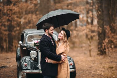 Gorgeous newlywed bride and groom posing in pine forest near retro car in their wedding day