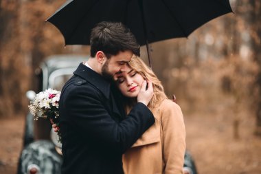 Gorgeous newlywed bride and groom posing in pine forest near retro car in their wedding day