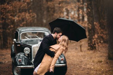 Gorgeous newlywed bride and groom posing in pine forest near retro car in their wedding day